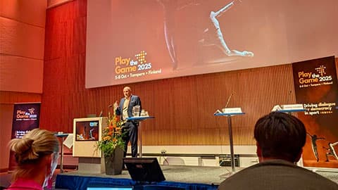 Alumnus Neal standing on stage with a screen in the background that reads Play the Game 2025. 5-8 October. Tampere. Finland.